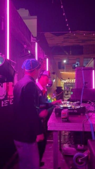 DJ in a beanie at the decks under pink neon.