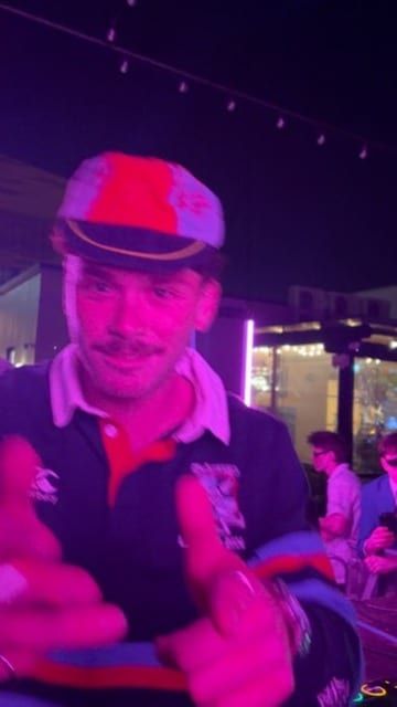 A grinning guy in a cap and rugby jersey, lit purple.