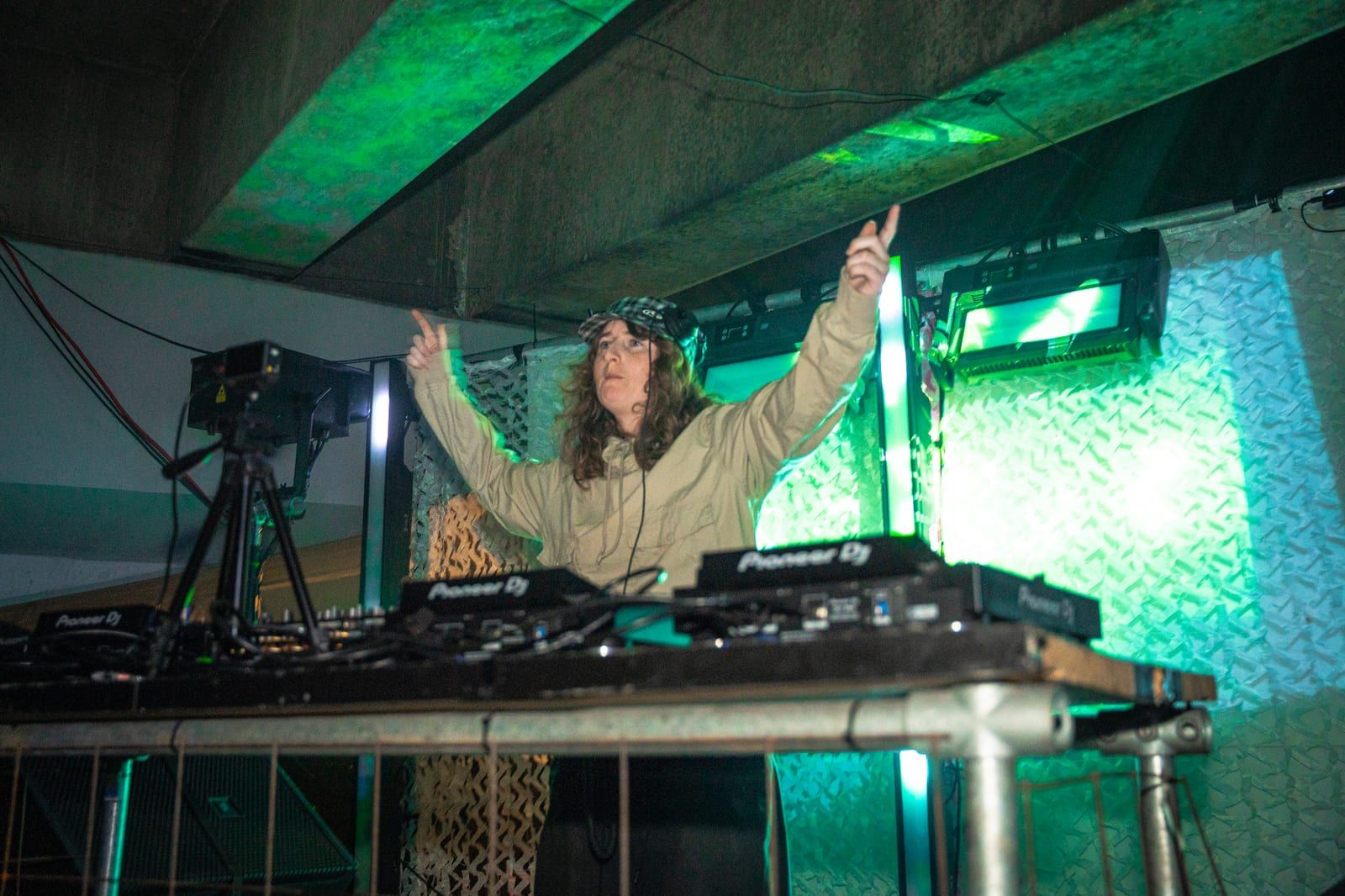 DJ with both arms in the air behind the decks, lit green under a concrete ceiling.