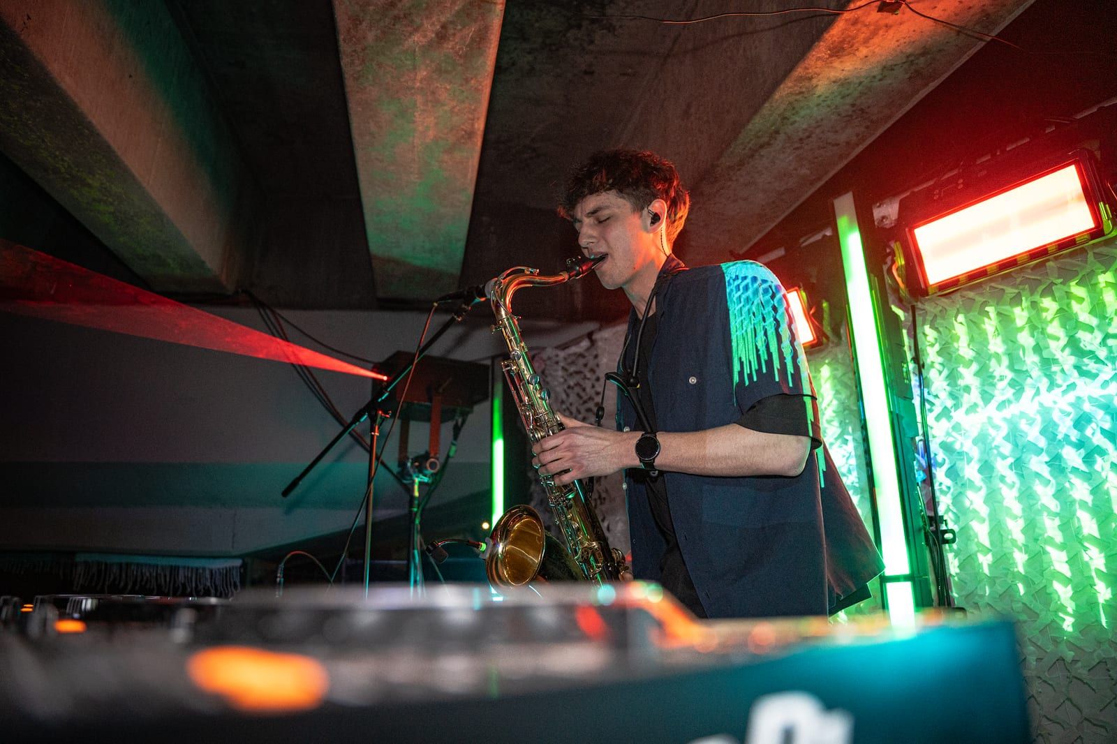 Saxophonist playing over the DJ booth, green and red light behind him.